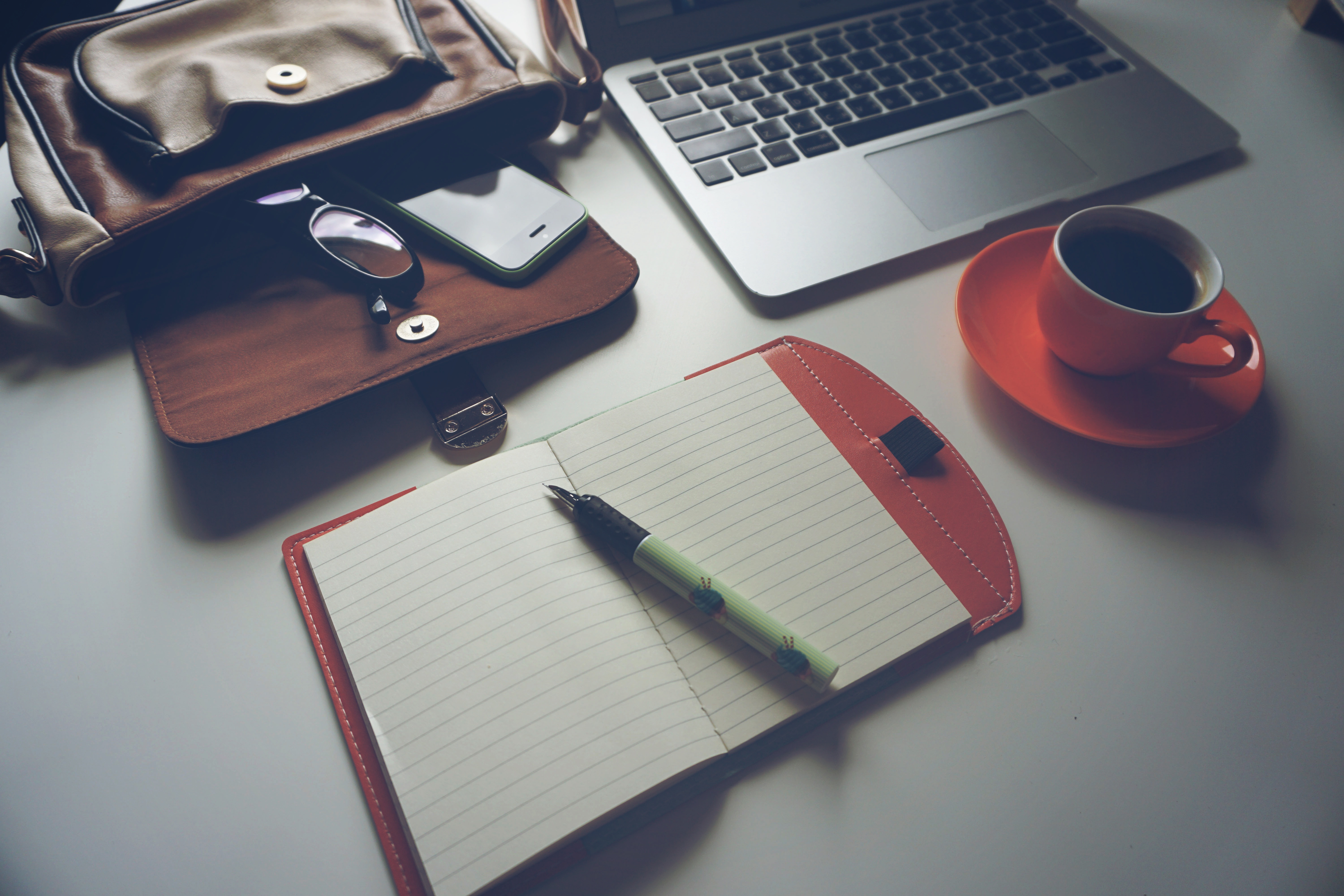 Open paper notebook with a fountain pen on it, beside a leather bag, a red mug of coffee, and a laptop